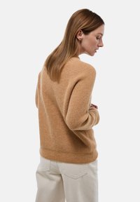 Pull en tricot beige à coupe décontractée, mettant en valeur une texture douce, des poignets côtelés et un col rond, associé à un pantalon de couleur claire.