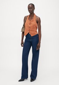 Oransje ermeløs vest med knapper, sammen med mørkeblå vide jeans og svarte spisse sko. Zebra-mønstredd håndveske over skulderen.