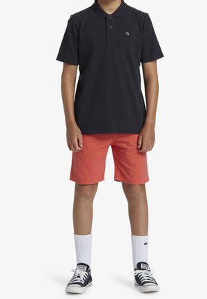 Garçon portant un polo noir, un short corail, des chaussettes blanches à hauteur de la cheville et des baskets Converse noires, debout devant un fond blanc.