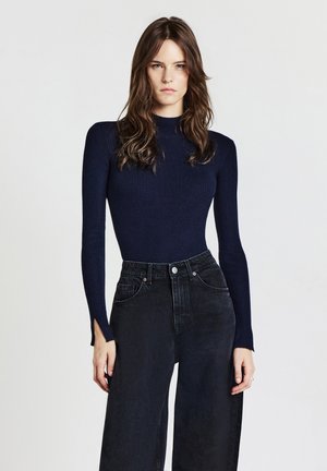 Maglietta a maniche lunghe a coste blu navy con colletto alto, abbinata a jeans neri a vita alta con gamba larga e spacchi sui lati delle maniche.