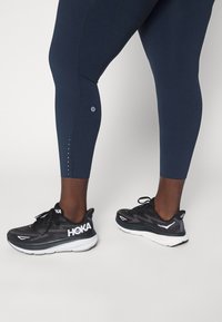 Leggings de color navy con una textura suave y pequeños puntos blancos a lo largo de la parte inferior de la pierna, combinados con zapatillas de correr negras que tienen un logo blanco.