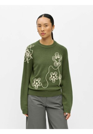 Femme portant un pull vert olive avec des motifs floraux abstraits blancs et un pantalon gris à jambes larges sur un fond blanc uni.