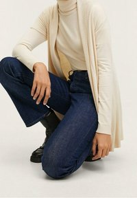 Personne à genoux portant un col roulé crème, un cardigan ouvert beige, un jean bleu foncé et des bottines noires sur un fond uni.