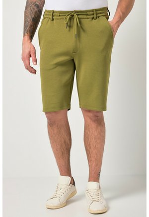Mand iført olivengrønne shorts med snører, hvide sneakers og en hvid skjorte, med en tatovering synlig på hans venstre underarm.