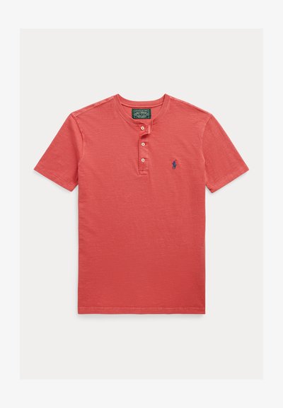 Korallfärgad pikétröja med korta ärmar, texturerad yta, tre knappar i knapplisten och en liten blå broderad logotyp på vänster bröst.