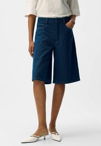 Persona che indossa pantaloni corti in denim scuro fino al ginocchio, una camicia bianca a maniche corte, un bracciale d'argento e scarpe slip-on bianche a tacco basso.