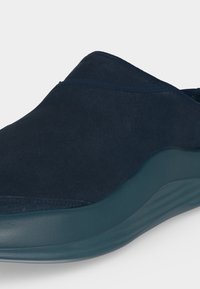 Σκούρο ναυτικό slip-on παπούτσι με λείο σουέντ άνω μέρος και παχύ, καμπυλωτό μπλε πάτο, εμφανίζεται σε κοντινή γωνιακή θέα.