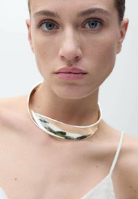 Söljmetallisk choker med en slät, böjd design. Reflekterande detaljer och en glansig yta kompletterar en ljus tygblus.