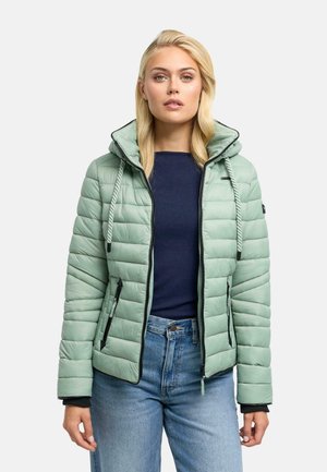 Frau mit blondem Haar, die eine hellgrüne gesteppte Jacke über einem dunkelblauen Top und blauen Jeans trägt, steht vor einem schlichten Hintergrund.