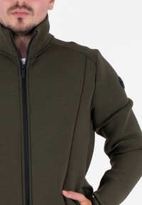 Giacca khaki verde con zip, colletto alto, texture liscia e zip nera. Presenta un accento rotondo nero sulla spalla.