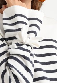 Top in tessuto bianco con righe orizzontali blu navy, caratterizzato da un dettaglio annodato sulla manica e una finitura morbida e testurizzata.