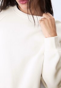 Bela sweatshirt s krojem okoli vratu in elastičnimi manšetami. Mehka tekstura tkanine, ki ima spuščena ramena in sproščen kroj. Brez vidnih vzorcev.