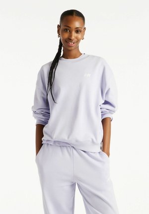Smilende kvinde med fletninger har lyse lilla sweatshirt og matchende sweatpants på, hænderne i lommerne, på ensfarvet hvid baggrund.