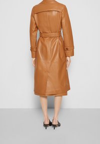 Manteau en cuir marron avec une ceinture à la taille, manches longues et boutons croisés. Il présente un pli à l'arrière pour faciliter les mouvements.