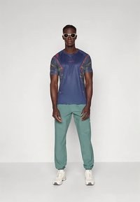 Vyras, vilkintis baltus saulės akinius, tamsiai mėlyną raštuotą sportinį marškinėlį, žalius sportinius kelnes ir baltus sportinius batelius, stovi prieš pilką fono.