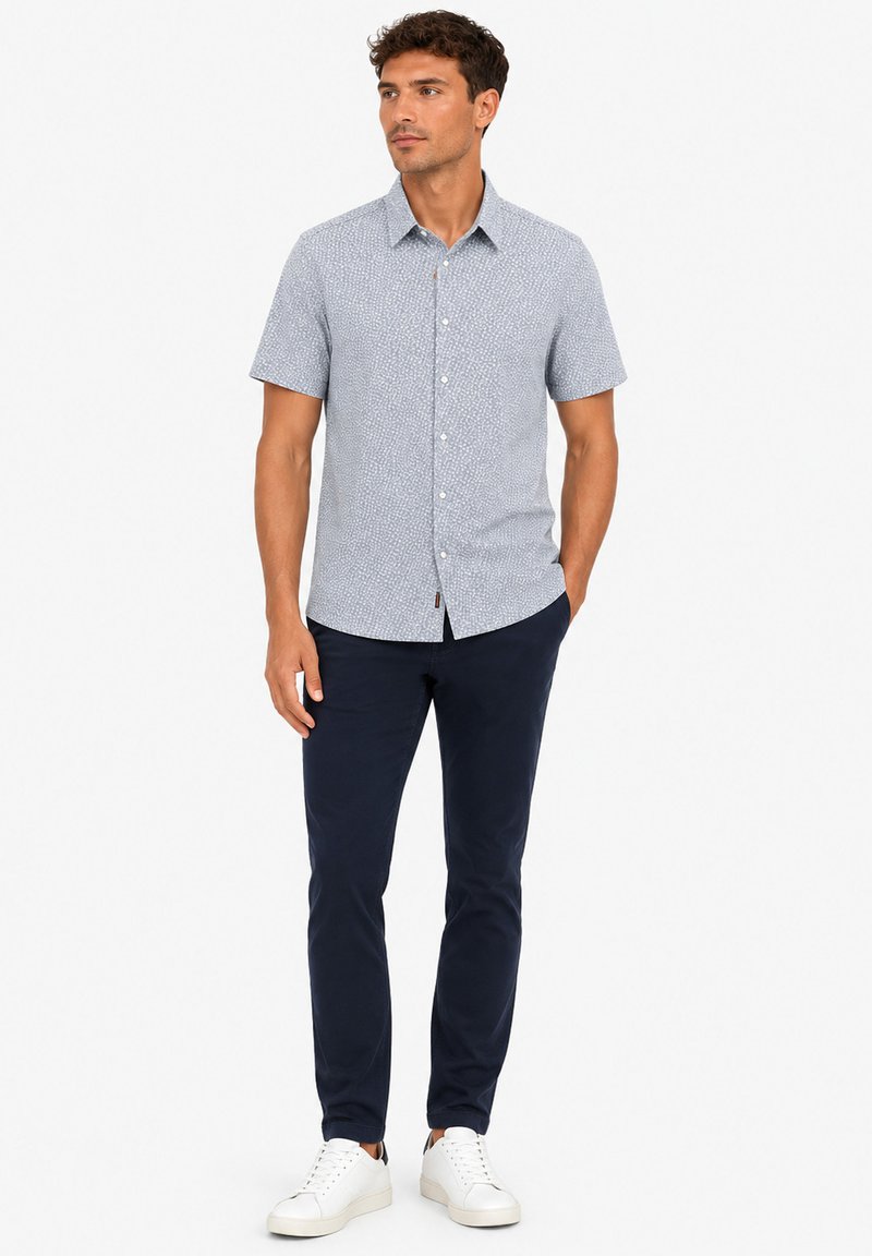 Homme debout portant une chemise légère à manches courtes à motifs, un pantalon slim foncé et des baskets blanches, mains détendues le long du corps.