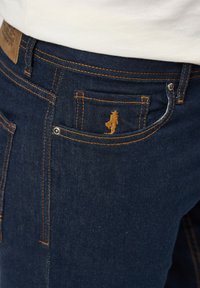 Jeans de mezclilla de color azul oscuro con costuras naranjas, un pequeño logo bordado en el bolsillo y remaches metálicos brillantes.