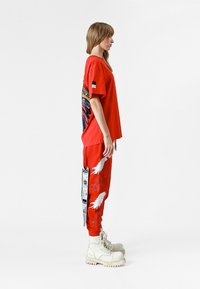 Camiseta roja de gran tamaño con un diseño de alas coloridas, combinada con pantalones de chándal rojos que presentan estampados de plumas blancas y una etiqueta negra. Botas de combate blancas.