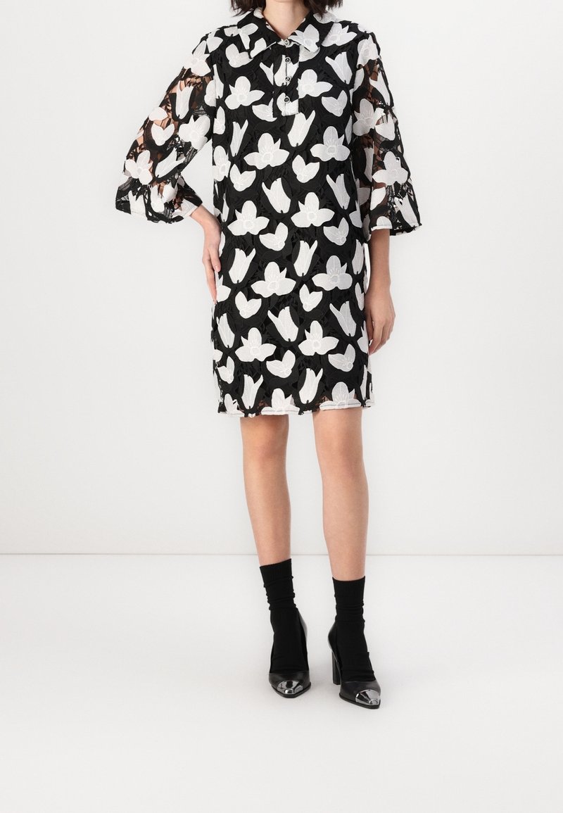 Femme portant une robe noire mi-longue avec des motifs blancs de papillons, manches transparentes 3/4, bottines chaussettes noires à bouts métalliques, debout, main sur la hanche.