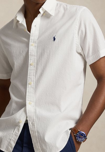 White short-sleeve shirt with a textured pattern, featuring a blue logo on the chest and yellow buttons. Worn with a blue watch and dark pants.