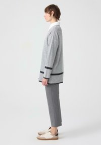 Grijze geribbelde cardigan met zwarte accenten, witte kraag, grijze op maat gemaakte broek en beige sneakers met een bruine zool, in zijprofiel.