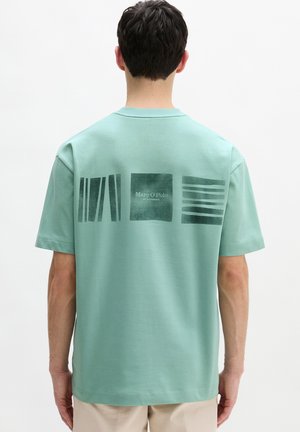 Mann trägt ein mintgrünes T-Shirt mit drei abstrakten, dunkelgrünen rechteckigen Designs und dem Schriftzug "Marc O'Polo" auf der Rückseite, steht vor einem weißen Hintergrund.
