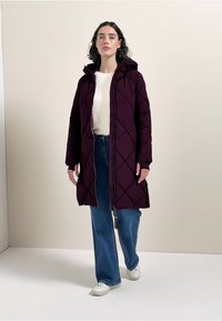 Cappotto lungo trapuntato bordeaux con cappuccio, caratterizzato da cuciture diagonali, zip frontale e tasche laterali, indossato sopra un maglione leggero e jeans blu.