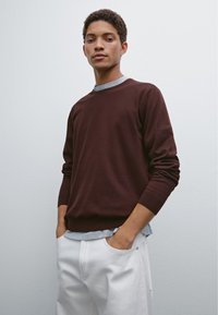 Maglione vinaccia con maniche lunghe, scollo a giro e orlo a costine, indossato sopra una maglietta grigia chiara. Abbinato a pantaloni bianchi.