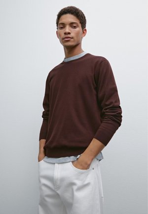 Maroon sweater with long sleeves, crew neckline, and a ribbed hem, layered over a light gray shirt. Paired with white trousers.
