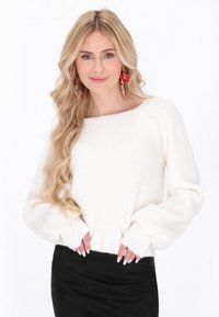 Pull en tricot blanc avec un large col et des poignets c�ôtelés, présentant un motif texturé. Porté avec une jupe noire, le modèle a de longs cheveux ondulés.