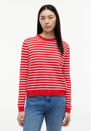 Femme portant un haut à manches longues rayé horizontalement rouge et blanc et un jean bleu, debout devant un fond blanc uni.