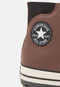 Bottine marron en cuir végan avec un panneau élastique noir, présentant un logo rond noir « Converse All Star » avec un motif d'étoile et une couture blanche.