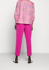 Blommig blus i rosa med blå och gula accenter, i kombination med livfulla rosa skräddarsydda byxor och vita slip-on skor. Slät tygtextur.