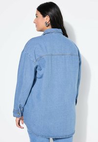 Lichtblauw denim shirt met een relaxte pasvorm, zichtbare stiksels, een kraag en knopenmanchetten, afgebeeld vanuit de achterkant tegen een witte achtergrond.