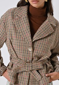 Cappotto a zig-zag intrecciato in marrone, beige e nero con vita modellata da cintura, grandi rever e due bottoni. Il tessuto ha una finitura testurizzata.