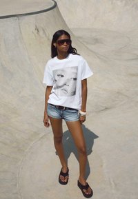 Hvid t-shirt med et sort-hvidt grafisk portræt. Blå denimshorts og sorte sandaler. Glat baggrund.