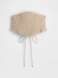Beige korsettopp med vertikale folder, som har en innsnevret midje og justerbare bånd bak med metalliske spisser. Glatt stoffstruktur.