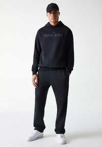 Felpa nera con cappuccio e logo grigio "NEW ERA", abbinata a pantaloni della tuta coordinati e scarpe bianche. Tessuto texture, vestibilità casual.