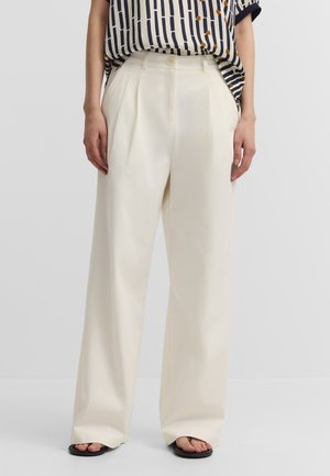 Weit geschnittene cremefarbene Hose mit vorderen Falten, kombiniert mit einem gestreiften kurzärmeligen Hemd und schwarzen offenen Sandalen.