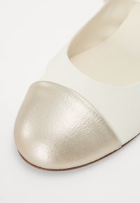 Ballerines couleur crème avec un embout brillant doré. Matière en cuir lisse, forme arrondie et détails sur les côtés ouverts.