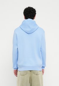 Ανοιχτό μπλε φούτερ με κουκούλα από απαλό υλικό· διαθέτει τσέπη καγκουρό και ριμπ μανσέτες και τελείωμα. Φοριέται με ανοιχτόχρωμο παντελόνι φόρμας.