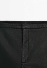 Gros plan sur un pantalon en cuir noir montrant la couture avant et la ceinture sur un fond blanc.