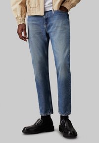 Ljust blå denimjeans med slim fit, med blekta områden och rak benform. Kombinerat med svarta loafers och en beige bomberjacka.