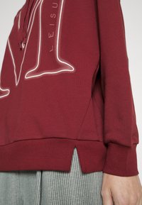 Gros plan d'une personne portant un sweat à capuche bordeaux avec un texte en contour blanc et des fentes latérales, associé à un pantalon gris à rayures fines.