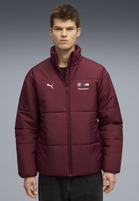 Veste rembourrée bourgogne avec un col haut, fermeture éclair sur le devant et logos de BMW et Puma. Présente une texture lisse et une coupe carrée.