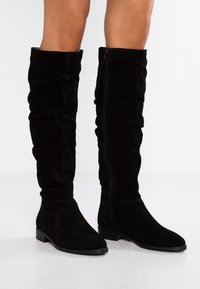 Knöchelhohe schwarze Wildlederstiefel mit einem taillierten Design, leichter Raffung oben und seitlichem Reißverschluss. Flache Sohle und glatte Textur.