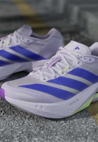 Λευκά και μωβ παπούτσια τρεξίματος Adidas με μπλε ρίγες και κείμενο "ADIZERO", τοποθετημένα σε τραχιά επιφάνεια πεζοδρομίου.