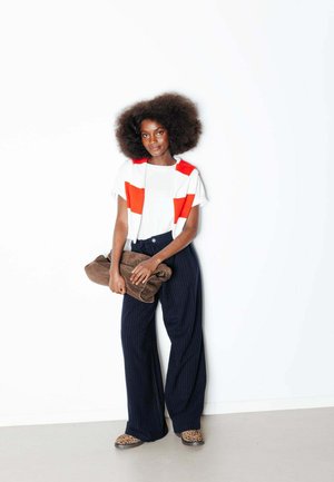 Jeune femme aux cheveux afro portant un haut blanc et rouge, un pantalon large à rayures bleu marine, des chaussures à imprimé léopard, tenant un manteau marron plié.