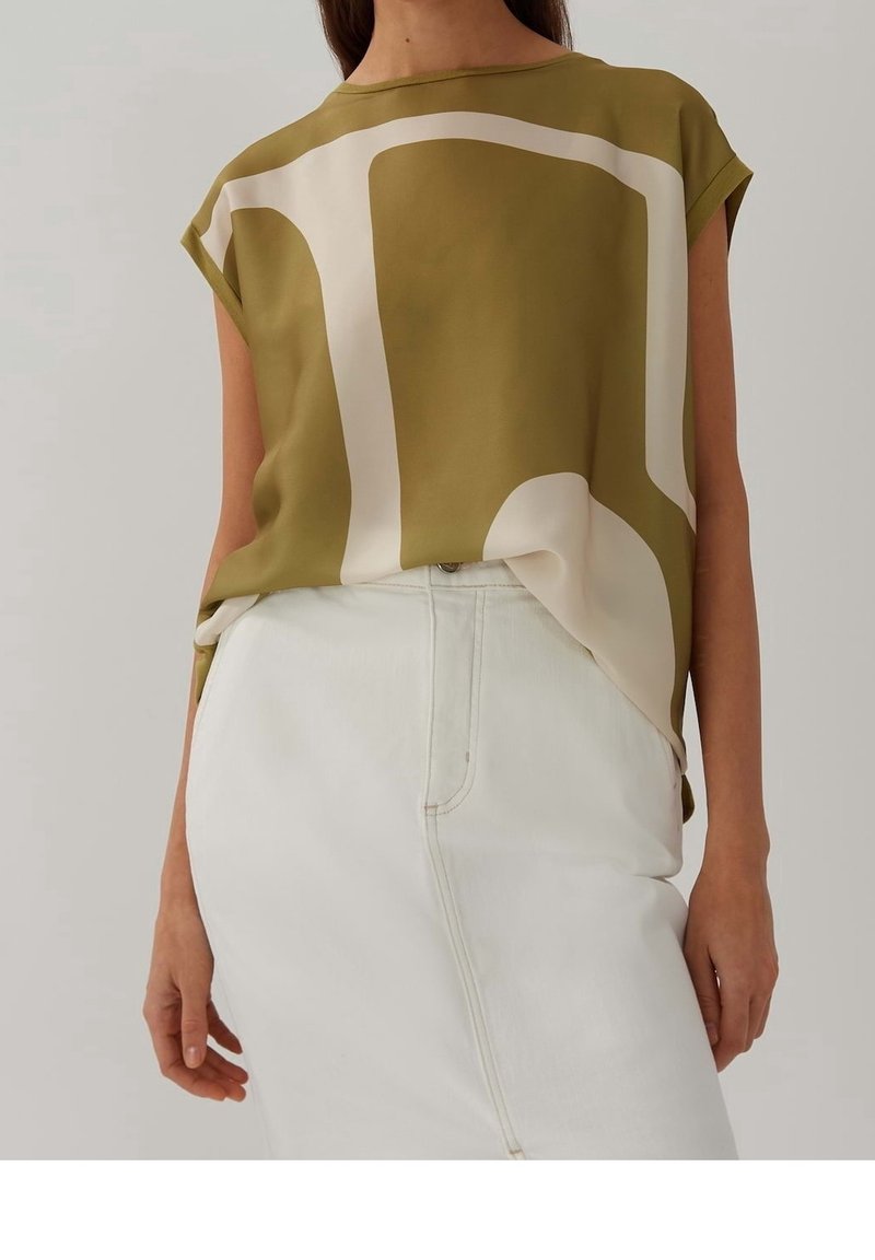Femme portant un haut sans manches vert olive avec un motif abstrait beige et une jupe blanche taille haute sur fond uni.