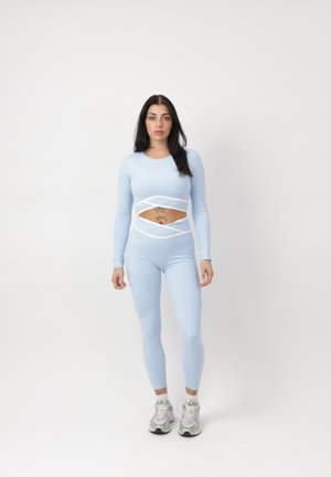 Woman with long dark hair wearing a light blue fitted two-piece outfit with cutouts and white sneakers standing against a plain background.